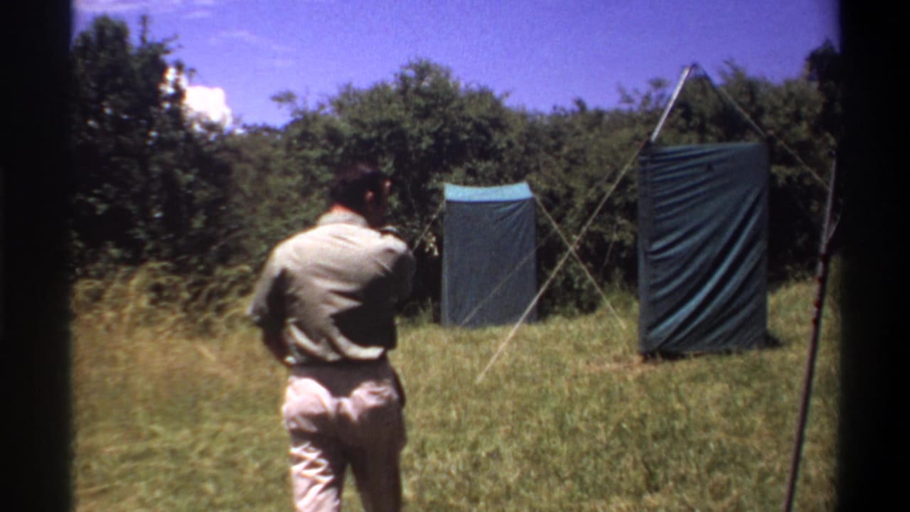 1969 Lagos home-movie frame with a rear view of a man nearing two tarp-covered structures in a sunlit grassy area.