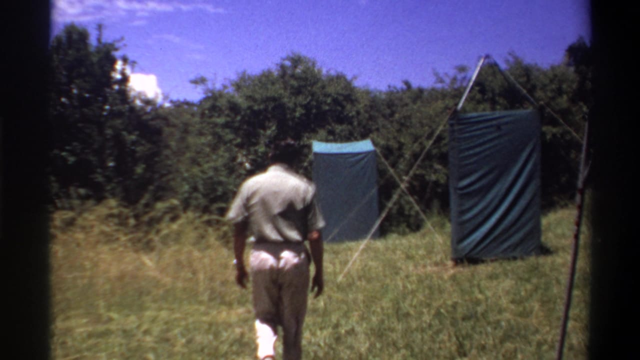 1969 Lagos frame showing the man farther into the field, with two blue-green tarp shelters, poles, and tree cover ahead.