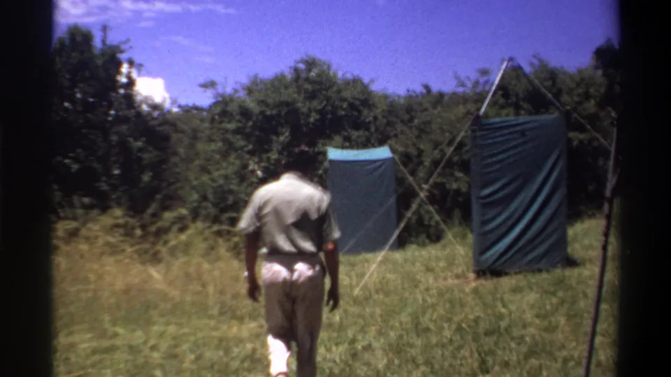 1969 Lagos frame showing the man farther into the field, with two blue-green tarp shelters, poles, and tree cover ahead.