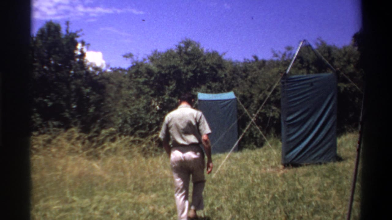 In supplied 1969 Lagos footage, a man in light clothing walks across grass toward blue tarp shelters near dense trees.