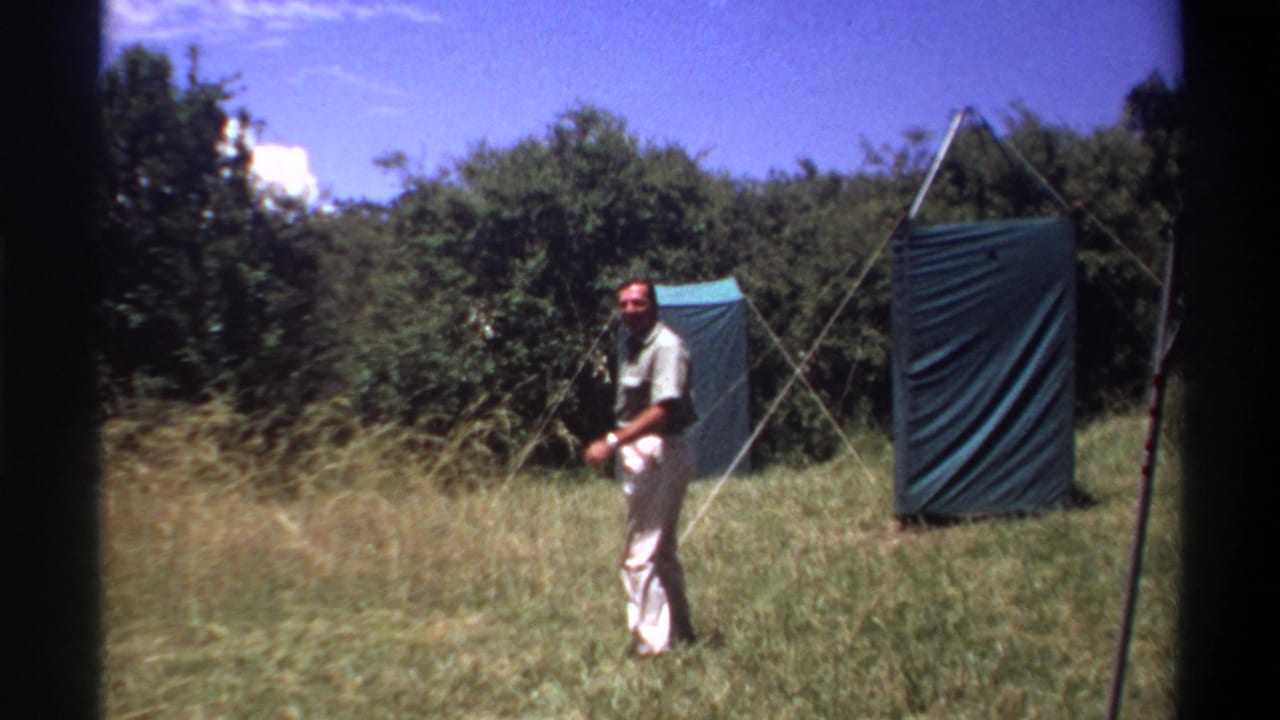The man turns back toward the camera in a Lagos grass field, with blue tarps, metal supports, trees, and sky behind him.