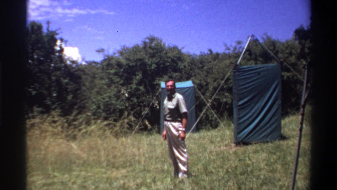 A smiling man stands facing the camera in the supplied Lagos 1969 scene, framed by tall grass and blue tarp panels.