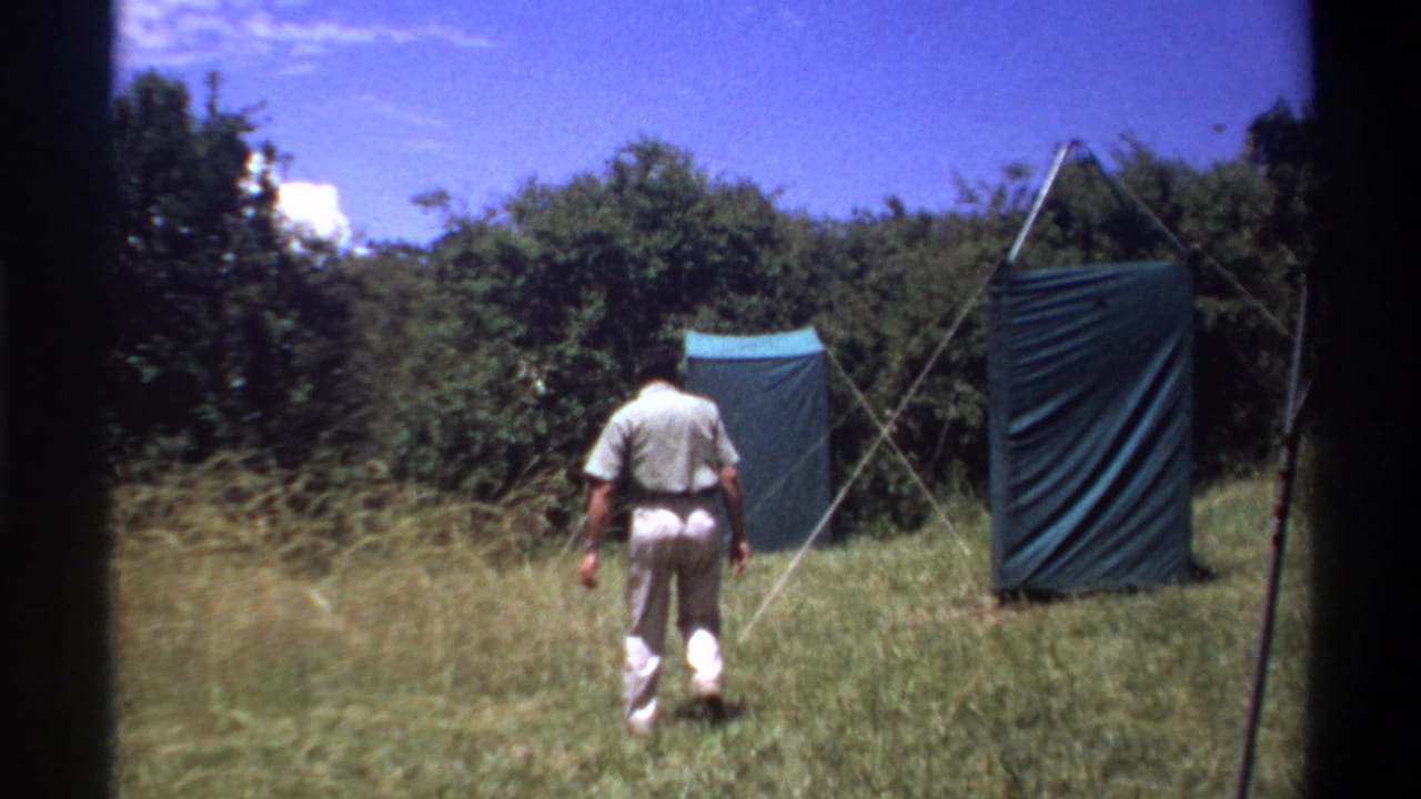 The man continues away across the Lagos grass field toward blue tarp shelters, with trees and bright sky in the background.