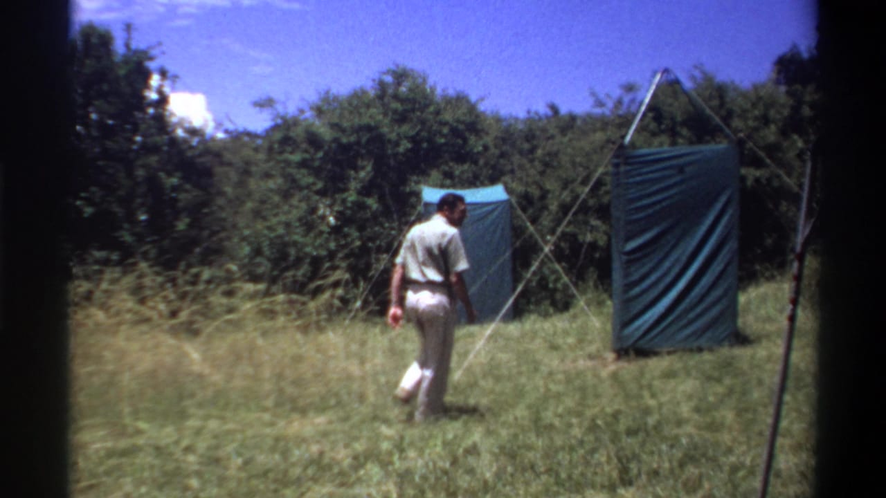 Man walking through a grassy clearing toward a green canvas tent among trees.