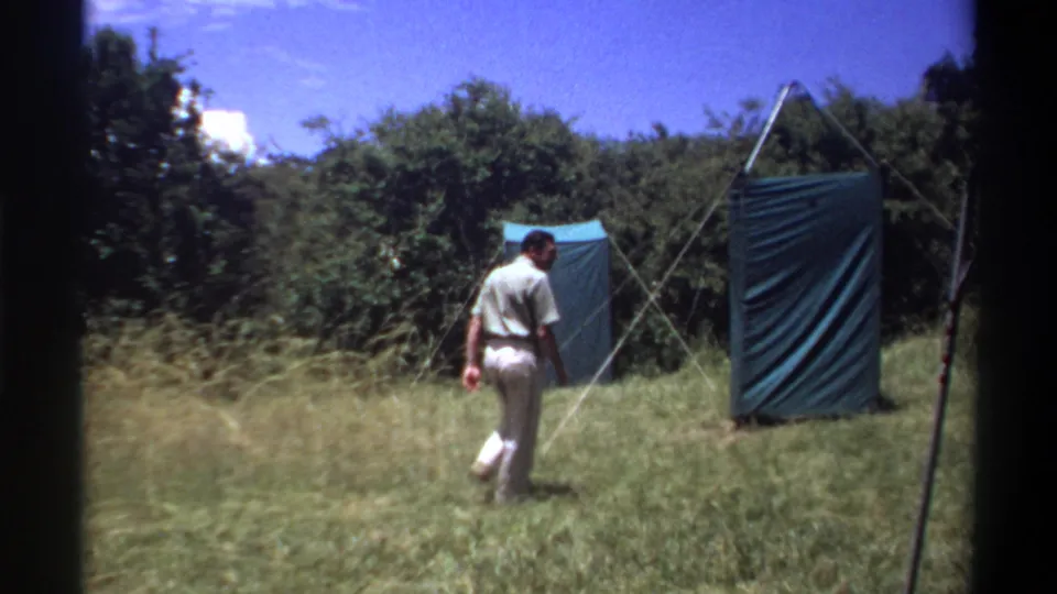 1969 Lagos home-movie frame showing a man in light clothing walking toward a green canvas tent set in tall grass and trees.