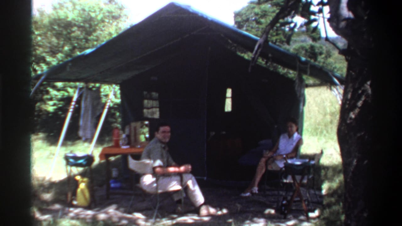 Two adults seated outside a large green tent with folding chairs and a small table.