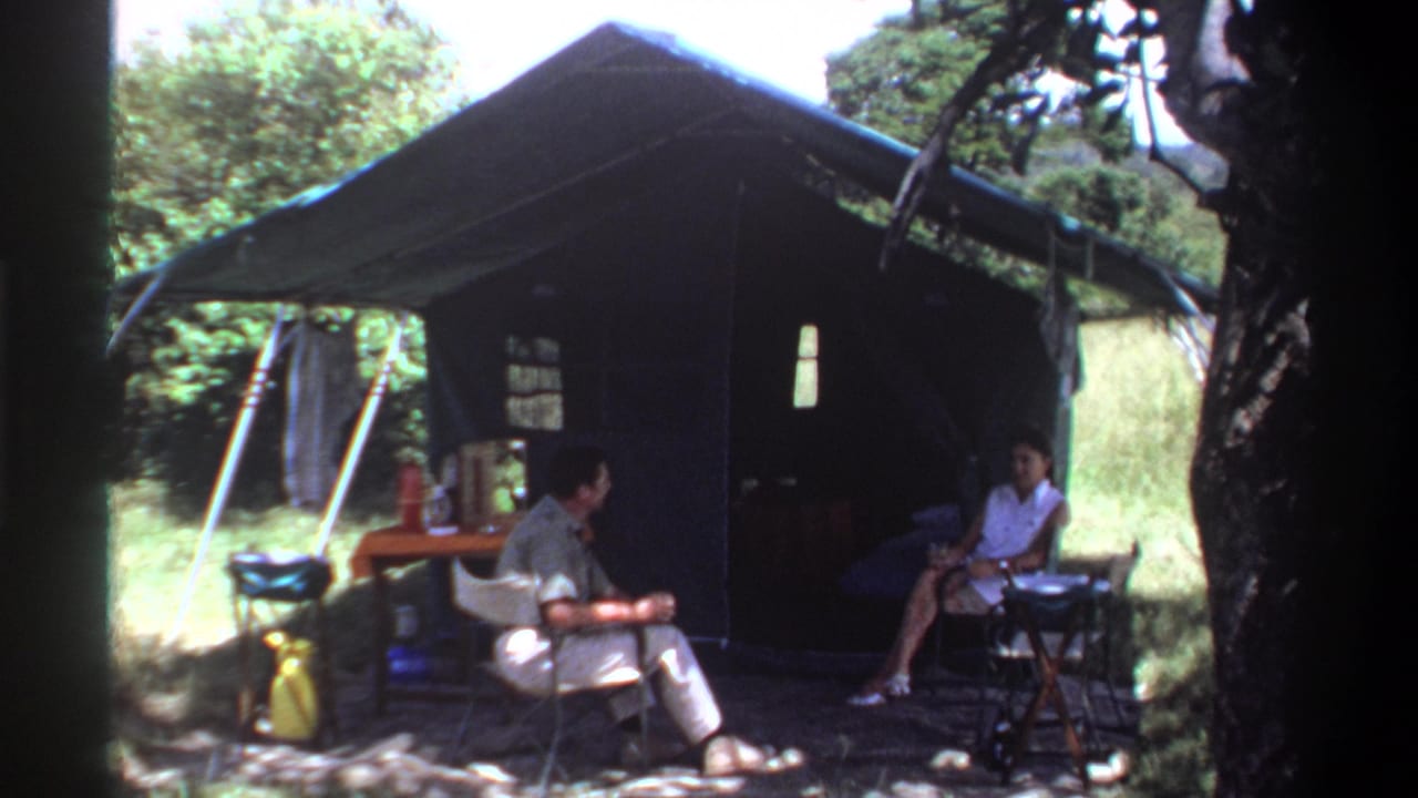 1969 Lagos home-movie frame showing seated adults at a campsite beside a large green tent under tree shade.