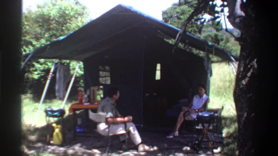 1969 Lagos home-movie frame showing seated adults at a campsite beside a large green tent under tree shade.