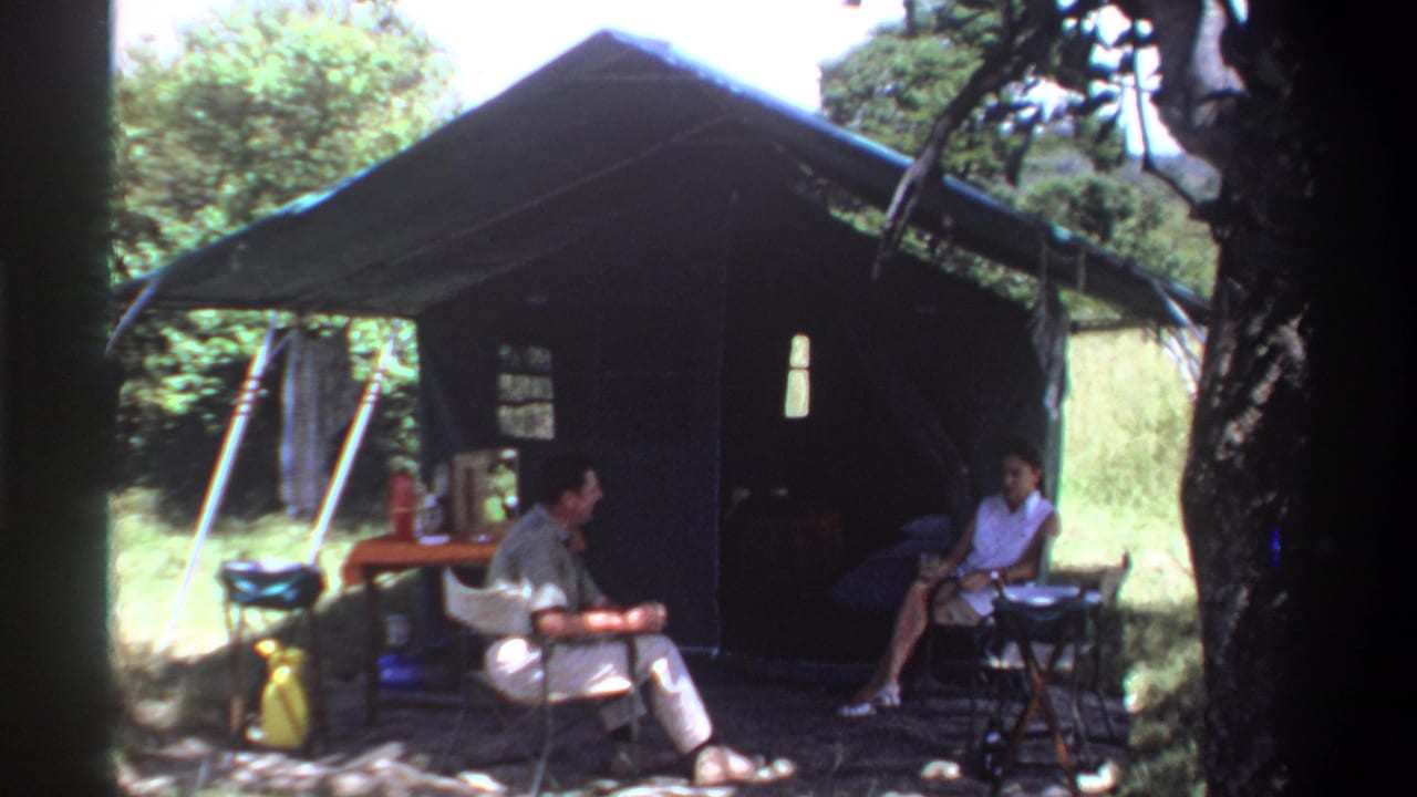 1969 Lagos home-movie frame showing two adults seated outside a canvas tent with a table and bottles nearby.