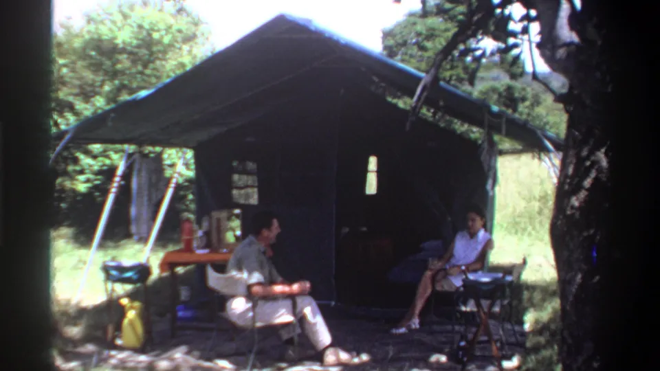1969 Lagos home-movie frame showing two adults seated outside a canvas tent with a table and bottles nearby.