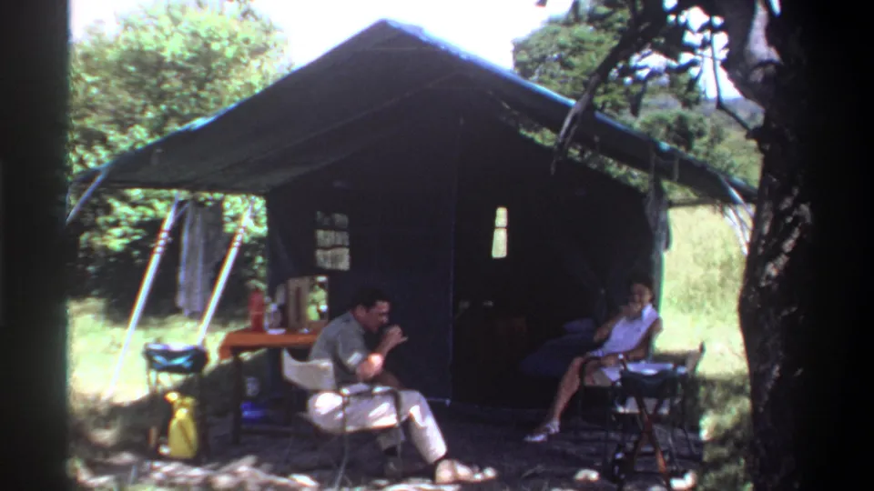 1969 Lagos campsite frame showing a seated man lifting a glass toward a woman outside a canvas tent.