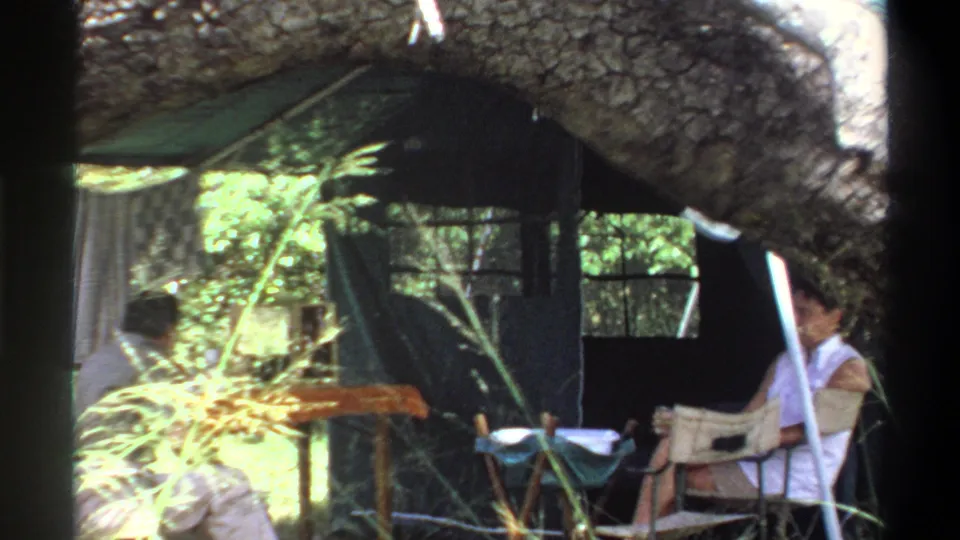 Adults sit near a canvas tent in a wooded Lagos-area campsite, filmed through branches in 1969 home-movie footage.
