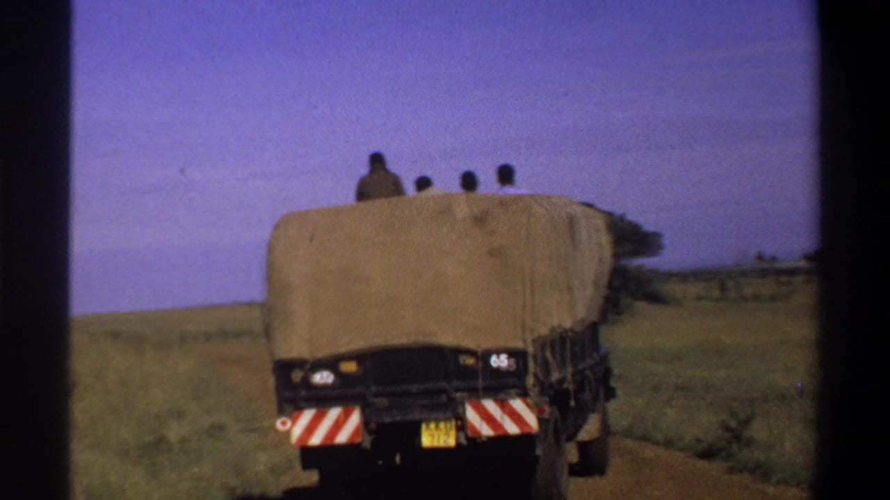 1969 Lagos, Nigeria frame of a canvas-covered truck moving away along a dirt road with passengers visible above the cover.