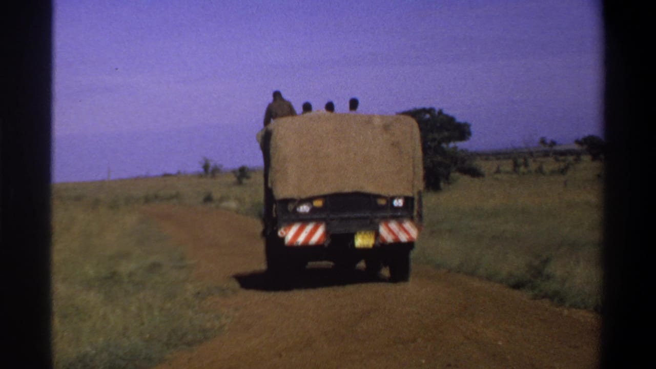 Wide 8mm view of a covered utility truck carrying passengers away along an unpaved road in the stated 1969 Lagos setting.
