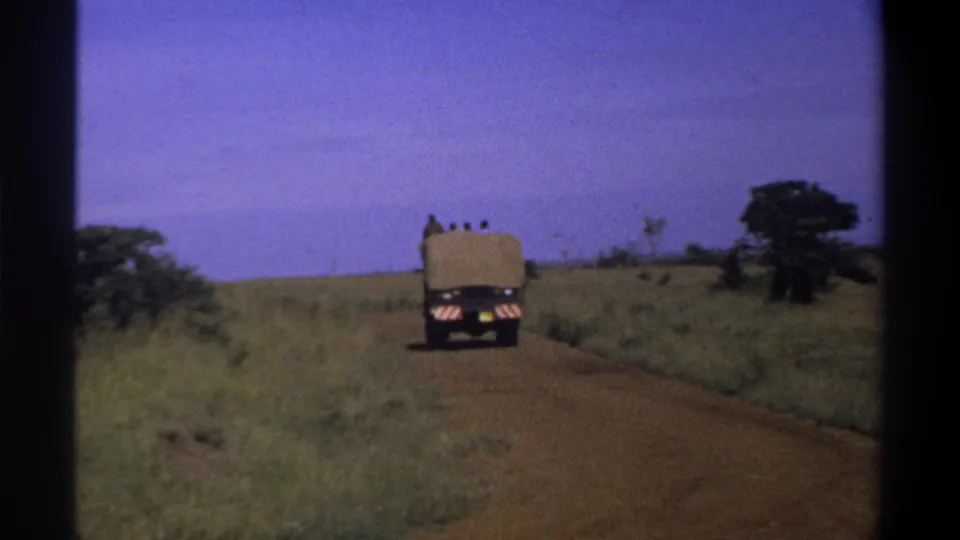 1969 Lagos, Nigeria 8mm frame showing the covered truck farther away on the reddish road through flat grassland.