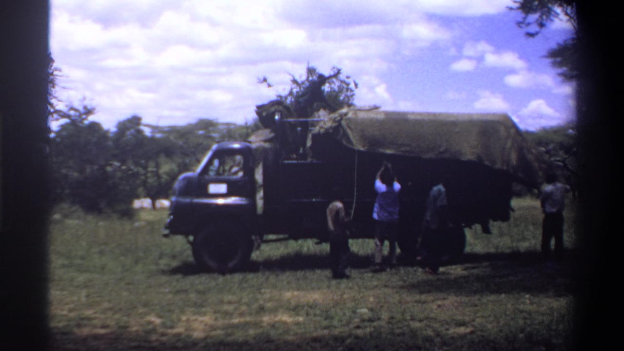 1969 Lagos-area home-movie frame showing men beside a dark tarp-covered truck in a grassy, tree-lined setting.