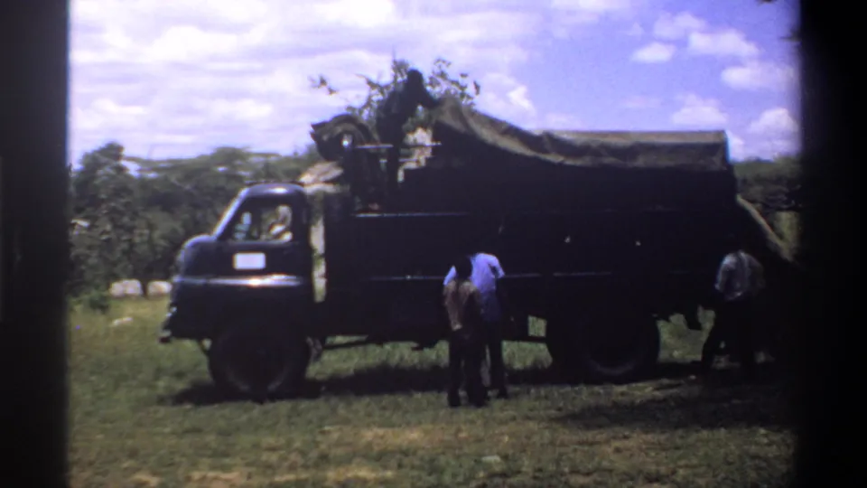 The final frame shows a dark covered truck and two men in the foreground, filmed in 1969 Lagos on grainy 8mm stock.