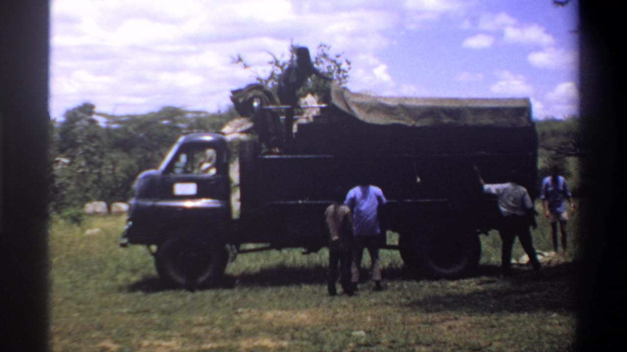 Five men stand around a covered truck in a grassy field under a bright sky.
