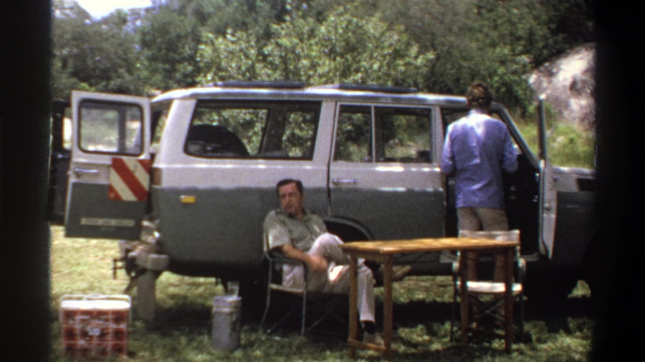 1969 Lagos home-movie frame showing one man seated smoking and another at an open station wagon door beside camp furniture.