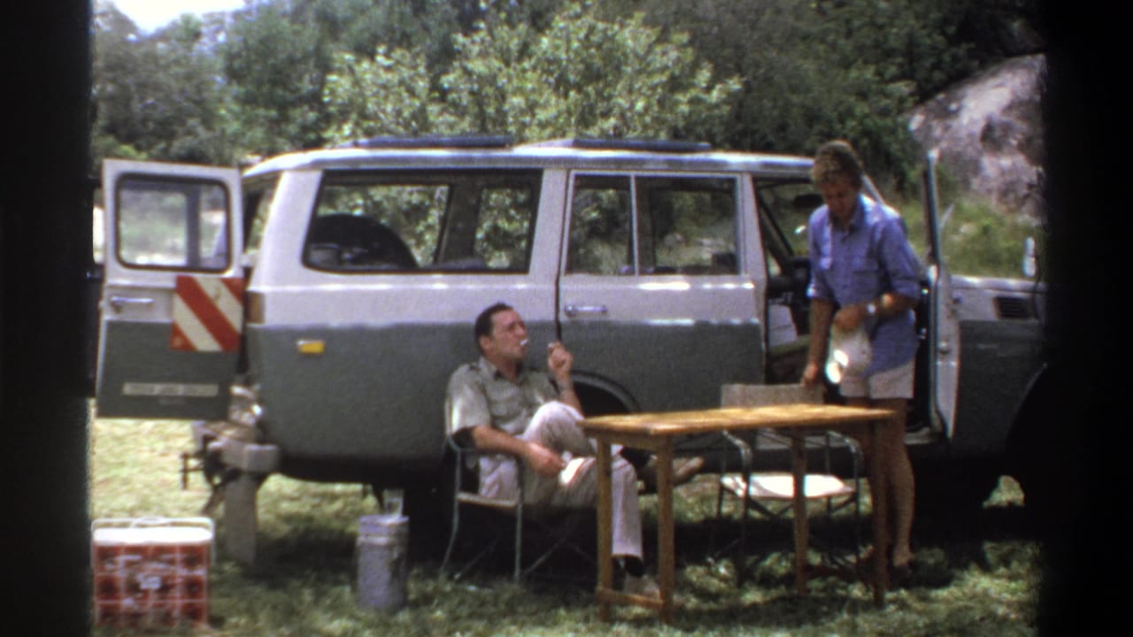 1969 Lagos home-movie frame showing a standing man holding a white bowl near camp furniture and a seated man by the station wagon.