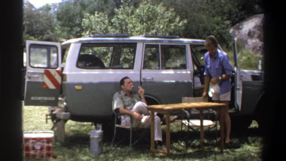 1969 Lagos home-movie frame showing a standing man holding a white bowl near camp furniture and a seated man by the station wagon.