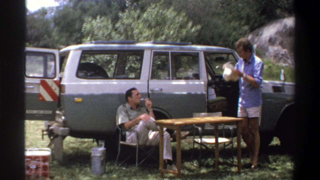 1969 Lagos campsite frame with a standing man holding a white bowl, a seated man smoking, and a two-tone station wagon behind them.