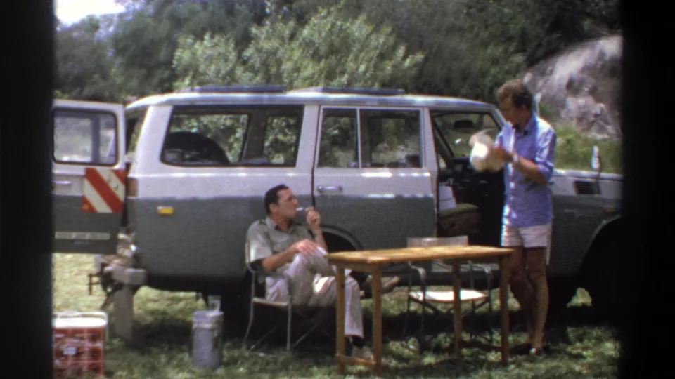 1969 Lagos campsite frame with a standing man holding a white bowl, a seated man smoking, and a two-tone station wagon behind them.