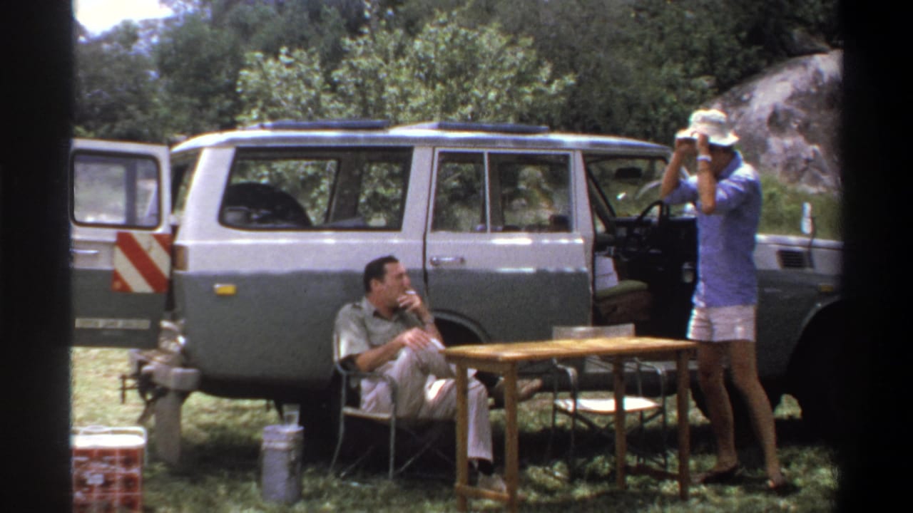 1969 Lagos home-movie frame showing two adult men near a two-tone station wagon, with folding chairs and a small outdoor table.