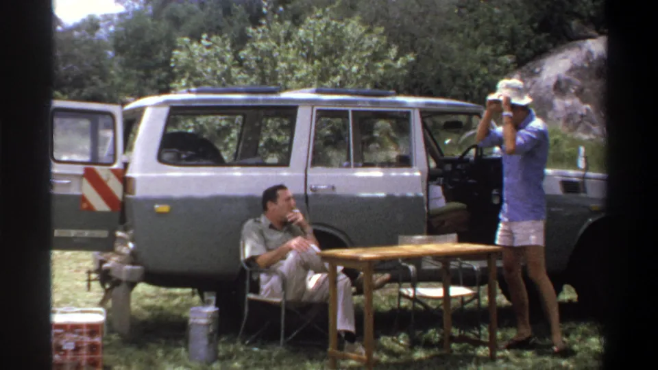 1969 Lagos home-movie frame showing two adult men near a two-tone station wagon, with folding chairs and a small outdoor table.