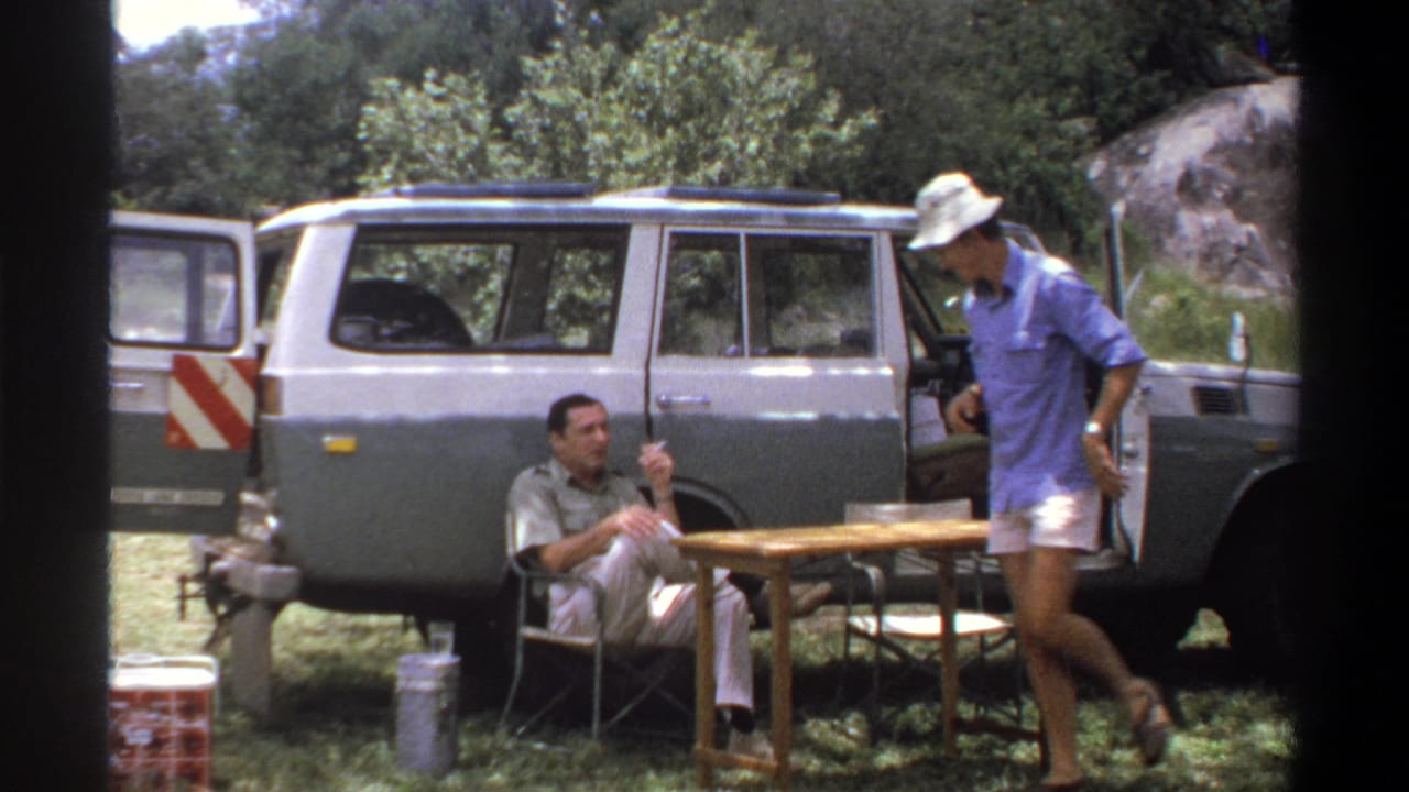 Man in a chair smokes while another man in a hat stands near an open station wagon door and table.