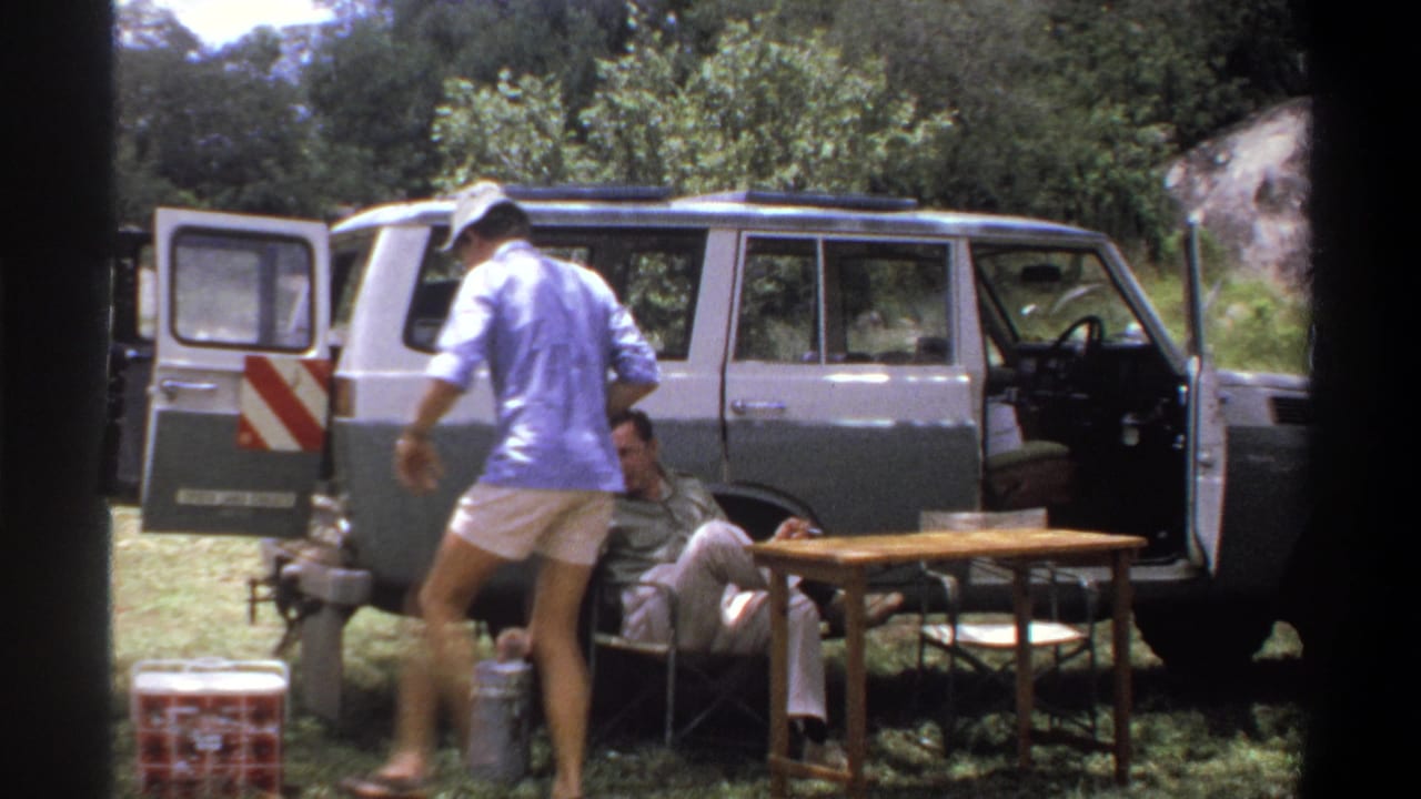 1969 Lagos home-movie frame showing a man in blue crossing the camp area beside a seated man, cooler, and open station wagon.