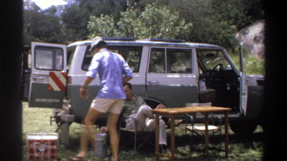 1969 Lagos home-movie frame showing a man in blue crossing the camp area beside a seated man, cooler, and open station wagon.
