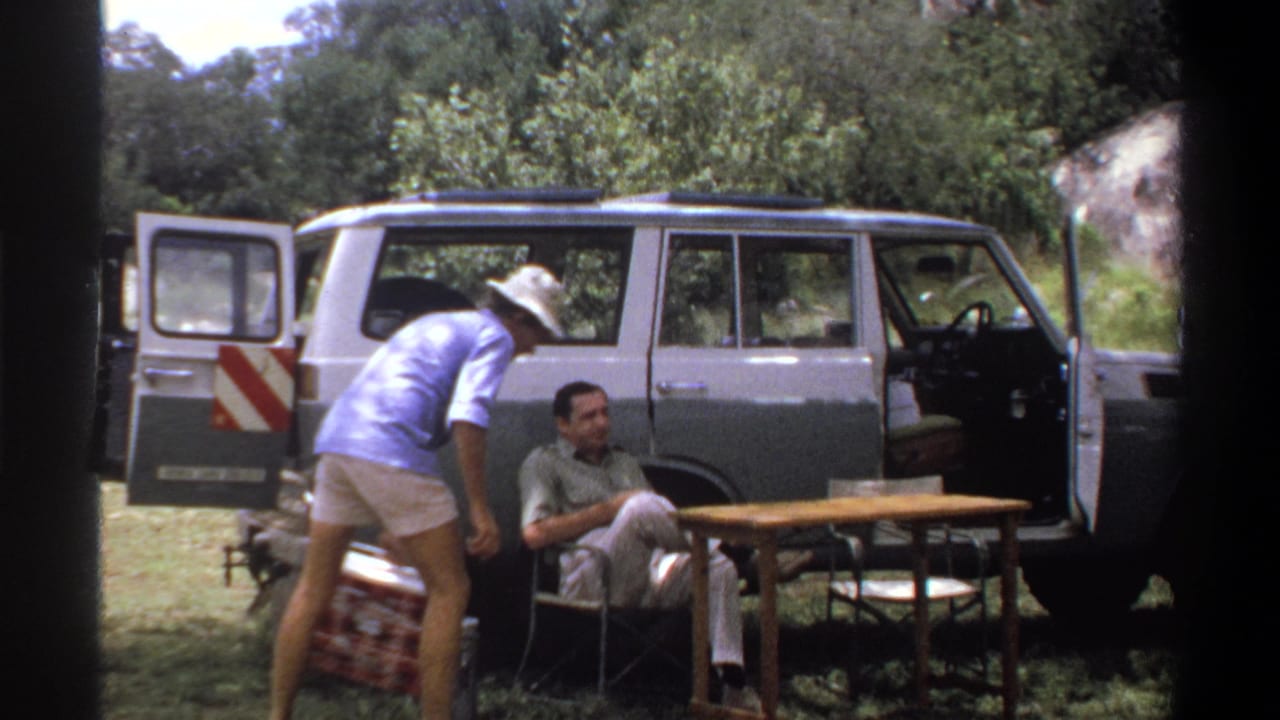 1969 Lagos frame with one man bending near a red cooler and another seated beside the table and open station wagon.