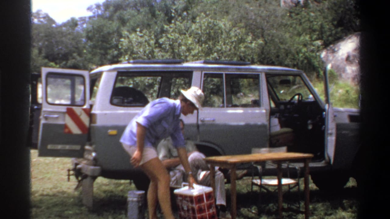 1969 Nigeria 8mm frame showing a man in a light shirt by a safari van with field supplies set out in grass.
