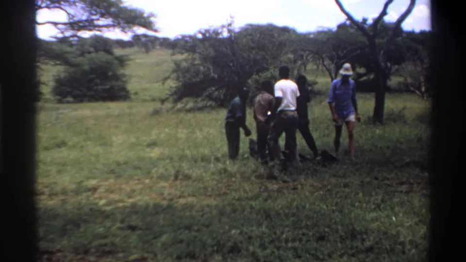 The 1969 Lagos home-movie frame shows several men standing close together around a dark object in tall grass.