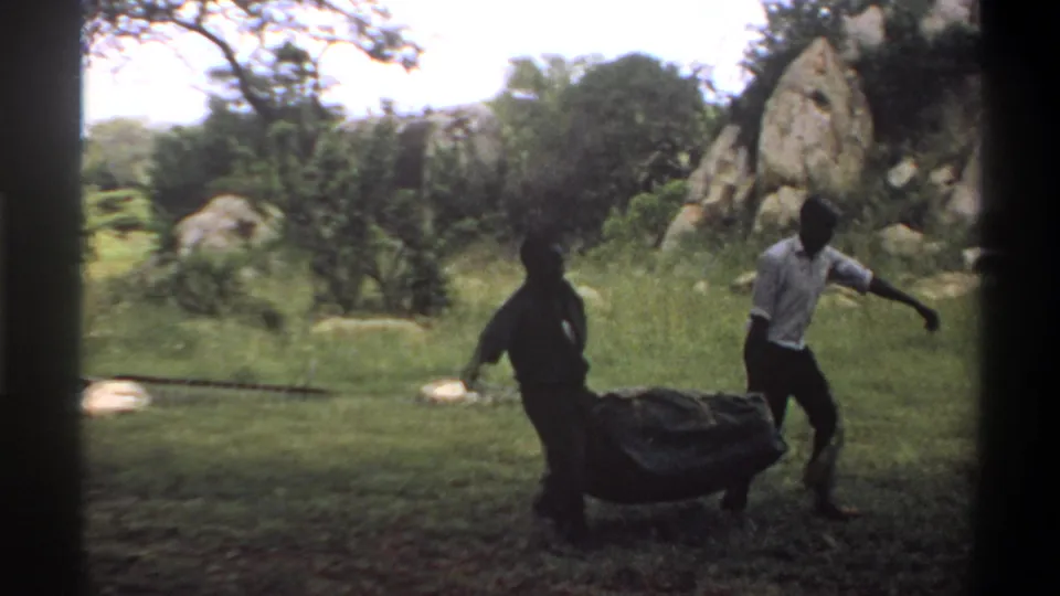 1969 Lagos, Nigeria 8mm frame showing men moving a covered bundle across grass near pale rocks and trees.