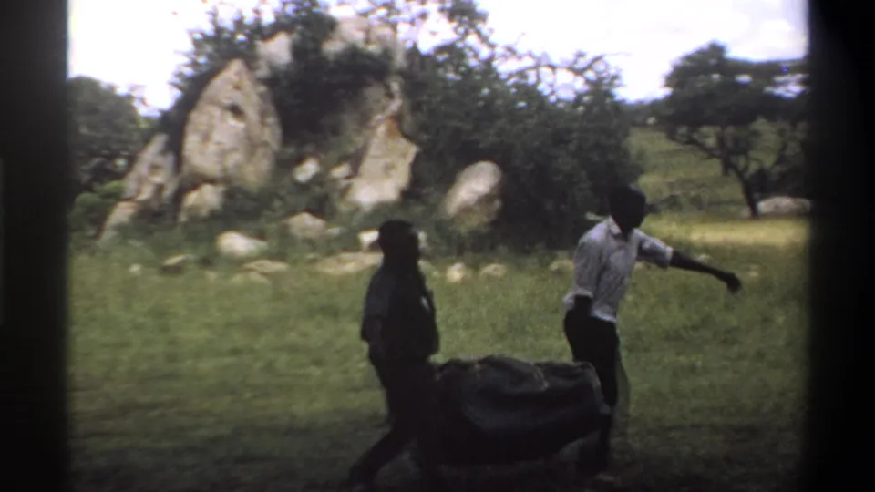 1969 Lagos, Nigeria frame showing two men carrying a covered load through grass with rocks and trees in the background.