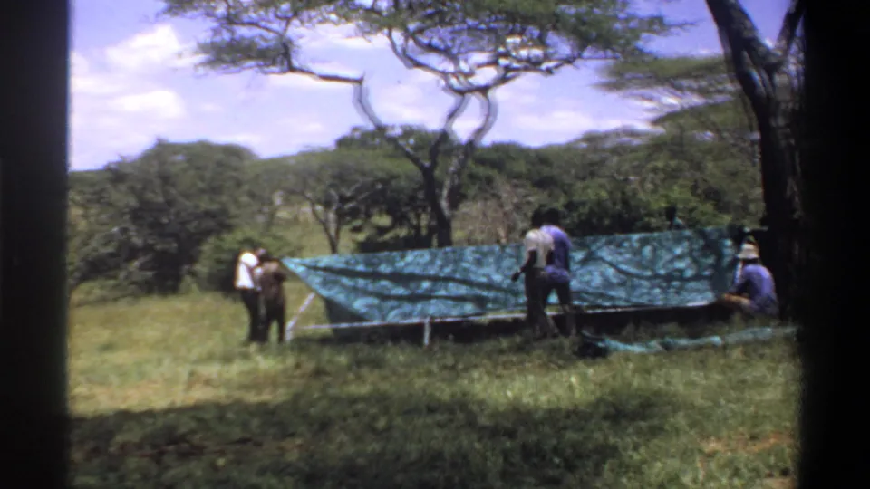 The final frame shows men beside a low tarp-covered structure in open grassland, with trees, hills, and 8mm frame edges visible.