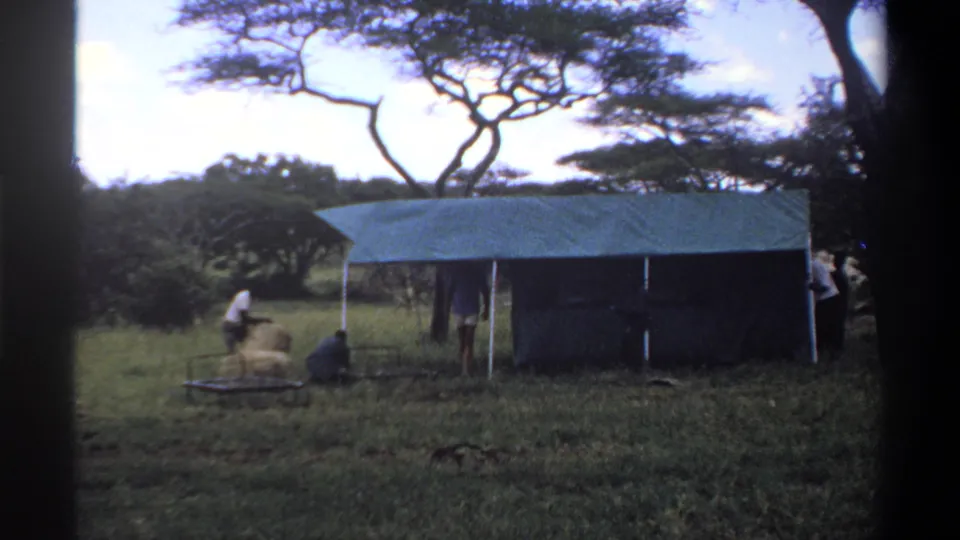 A person handles material near a low frame while others stand by a canvas shelter in 1969 Lagos-area footage.