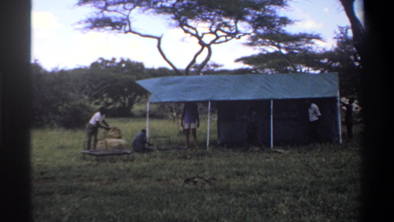 1969 Lagos, Nigeria 8mm frame showing men near a green tarp campsite in a grassy clearing with trees in the background.