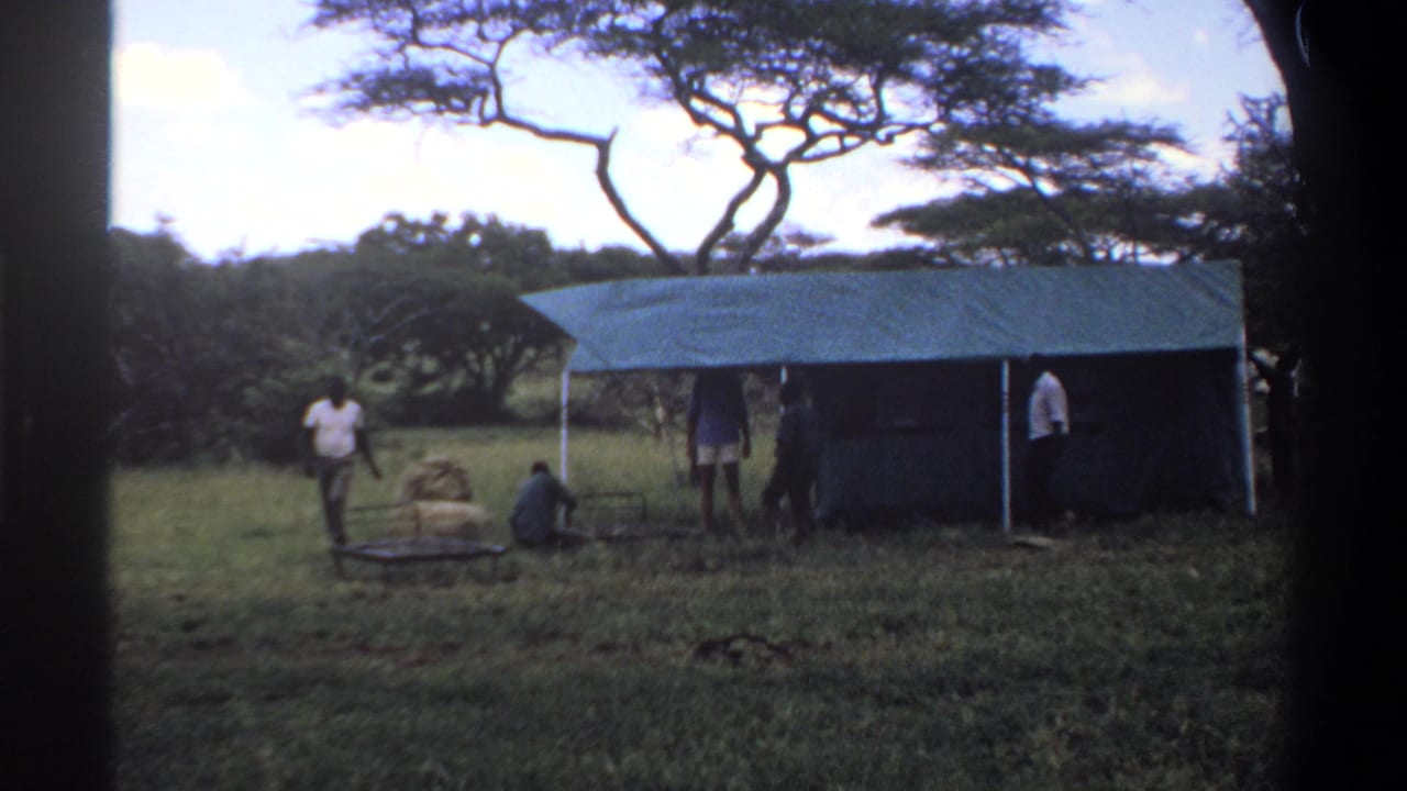 1969 Lagos, Nigeria 8mm frame with men around a green tarp shelter, one near bundled supplies and a metal frame in the grass.