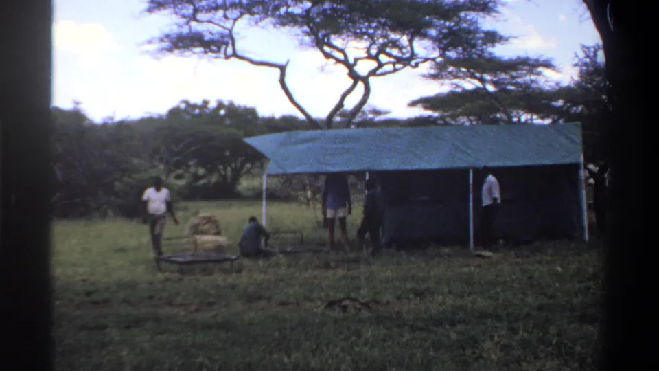 1969 Lagos, Nigeria 8mm frame with men around a green tarp shelter, one near bundled supplies and a metal frame in the grass.