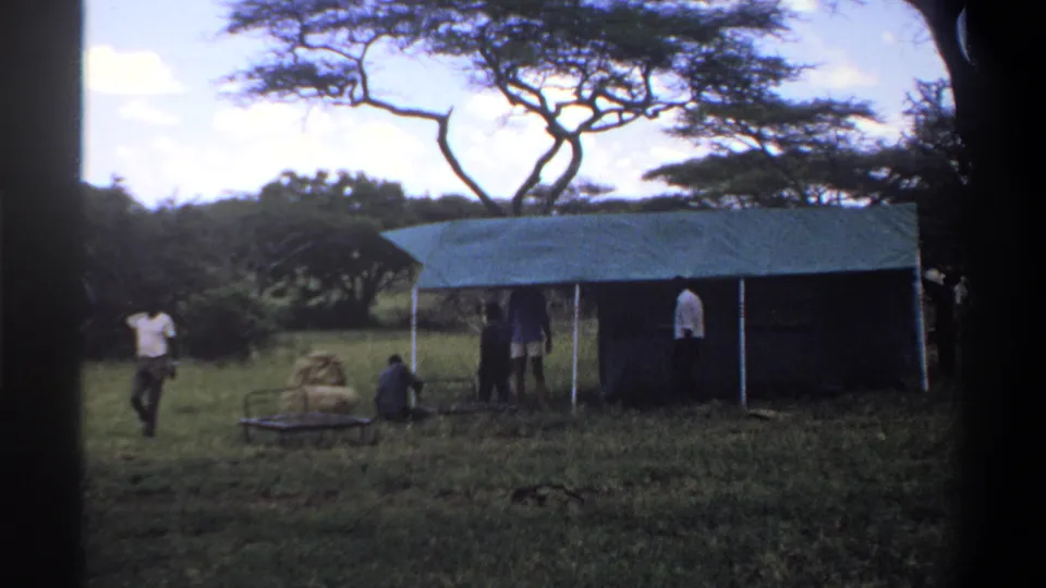 1969 Lagos, Nigeria 8mm frame showing a man walking past a camp area while others stand or sit near a green tarp shelter.