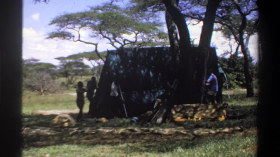 1969 Nigeria home-movie frame showing several people clustered near a large canvas tent in a grassy, wooded clearing.