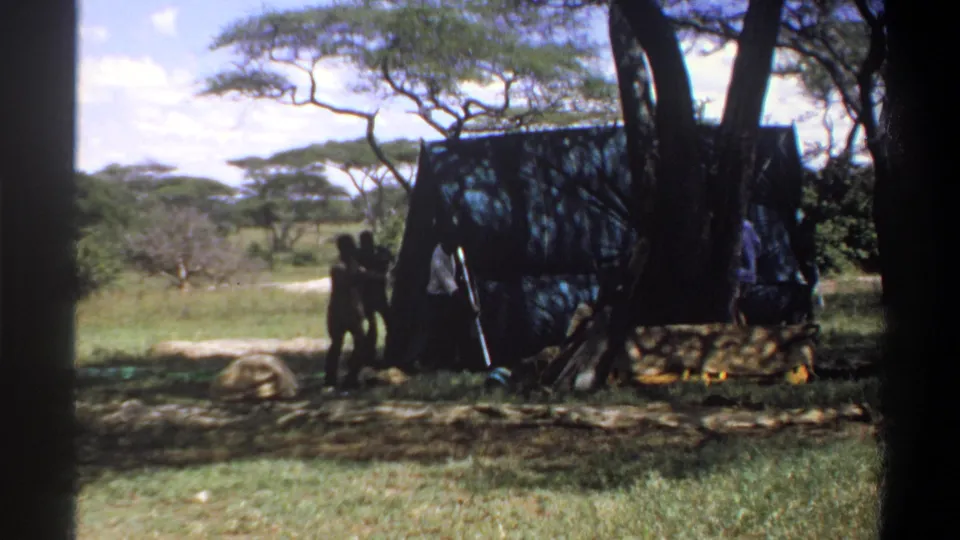 1969 Nigeria frame showing people beside a dark canvas tent, with grass, trees, and camp bundles visible in the clearing.