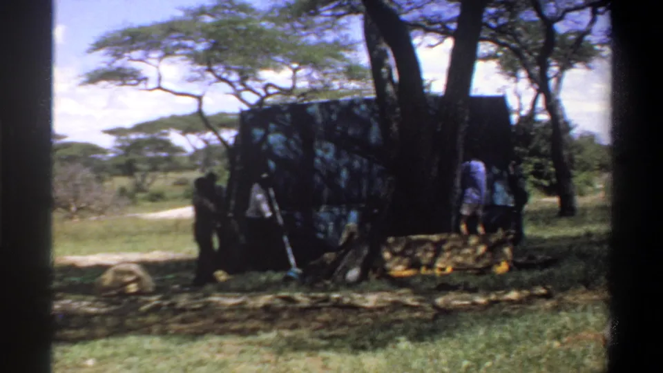 1969 Nigeria 8mm frame showing people working around a dark tent or tarp structure beneath trees in bright daylight.