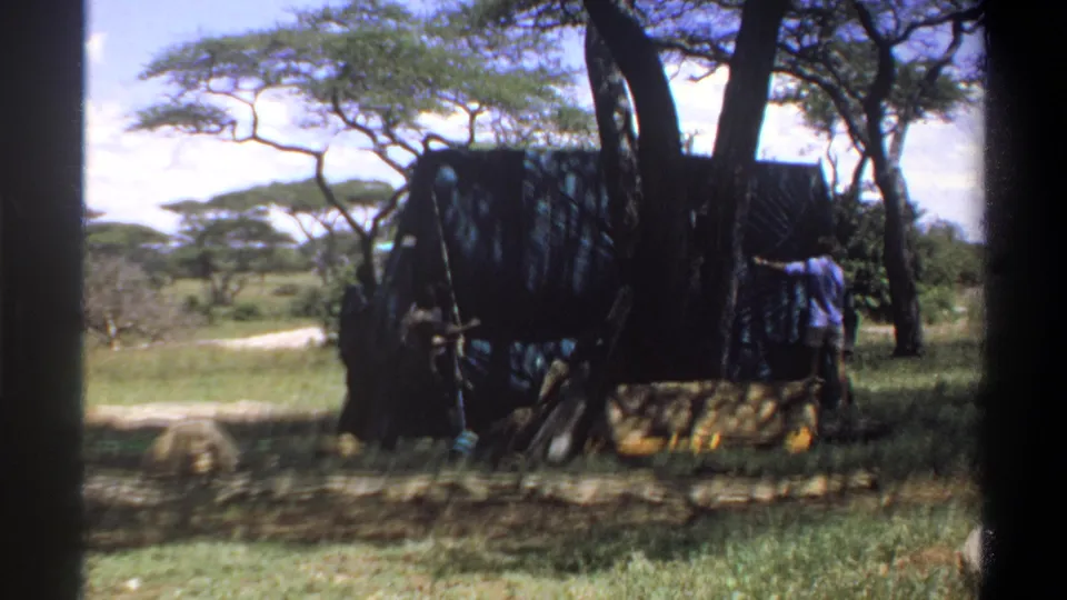 1969 Nigeria 8mm frame showing a large dark temporary structure under trees, with people positioned at its sides.