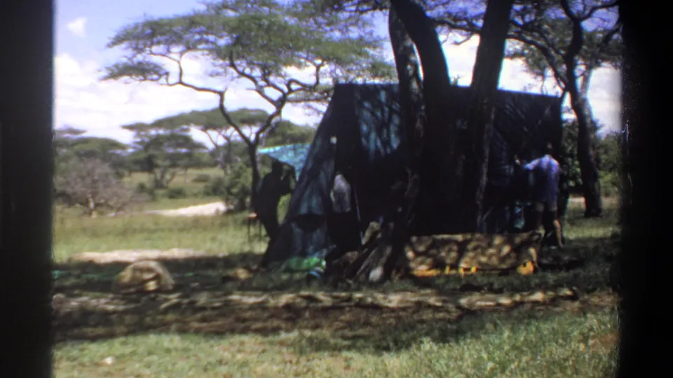 1969 Nigeria home-movie frame showing people beside a dark tent or tarp, with trees, grass, and a dirt path behind them.