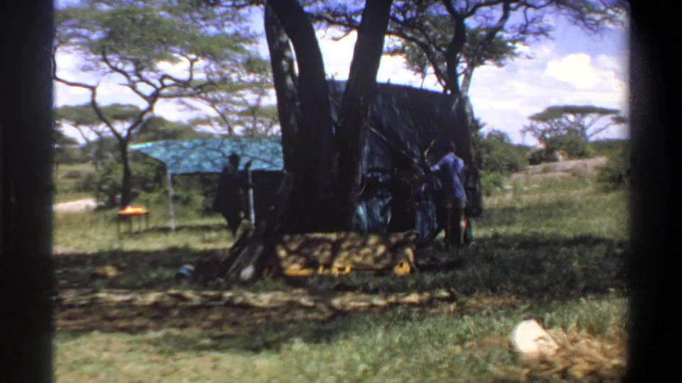 1969 Lagos, Nigeria frame with adults positioned around a dark temporary shelter in a grassy, tree-dotted outdoor setting.