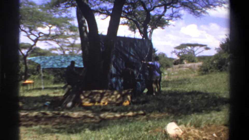 1969 Lagos, Nigeria home-movie frame with a person beside a tarp shelter, trees, grass, and a blue-roofed structure in the background.